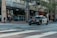 A police car with lights flashing is parked on a city street. Several pedestrians are standing on a sidewalk near an intersection, next to a building with multiple AT&T banners. Trees line the street and a one-way sign is visible.
