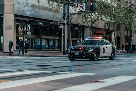 A police car with lights flashing is parked on a city street. Several pedestrians are standing on a sidewalk near an intersection, next to a building with multiple AT&T banners. Trees line the street and a one-way sign is visible.