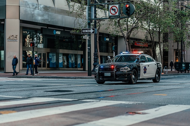 A police car with lights flashing is parked on a city street. Several pedestrians are standing on a sidewalk near an intersection, next to a building with multiple AT&T banners. Trees line the street and a one-way sign is visible.