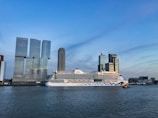 A large cruise ship is docked near a city skyline. The ship has distinctive artwork on its hull. Behind the ship, modern skyscrapers stand tall with a clear blue sky in the background. The water in the foreground reflects the light.