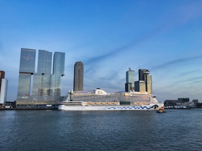 A large cruise ship is docked near a city skyline. The ship has distinctive artwork on its hull. Behind the ship, modern skyscrapers stand tall with a clear blue sky in the background. The water in the foreground reflects the light.