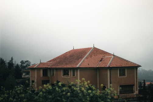A large, two-story house with a reddish-brown tiled roof is surrounded by fog and greenery. The structure has multiple windows and slightly muted yellowish walls. The atmosphere is misty, with trees visible in the background, adding to a serene and secluded ambiance.