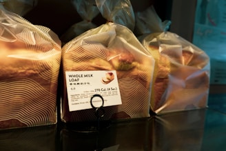Several loaves of bread are wrapped in translucent plastic bags, each with a printed label showing nutritional information and the name 'Whole Milk Loaf'. The bread is organized on a reflective surface, and the packaging has a modern design with wavy lines.