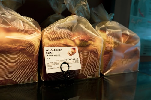 Several loaves of bread are wrapped in translucent plastic bags, each with a printed label showing nutritional information and the name 'Whole Milk Loaf'. The bread is organized on a reflective surface, and the packaging has a modern design with wavy lines.