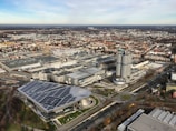 An aerial view of an urban industrial area featuring large factory buildings and office structures. In the foreground, a prominent building with a modern roof design is visible next to a circular tower complex with distinct round sections. Several roads and smaller buildings surround the area, indicating an industrial or business district. The scenery extends into a distant landscape with a blend of urban and natural elements.