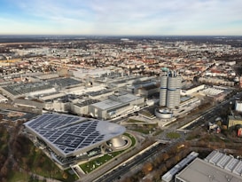 An aerial view of an urban industrial area featuring large factory buildings and office structures. In the foreground, a prominent building with a modern roof design is visible next to a circular tower complex with distinct round sections. Several roads and smaller buildings surround the area, indicating an industrial or business district. The scenery extends into a distant landscape with a blend of urban and natural elements.
