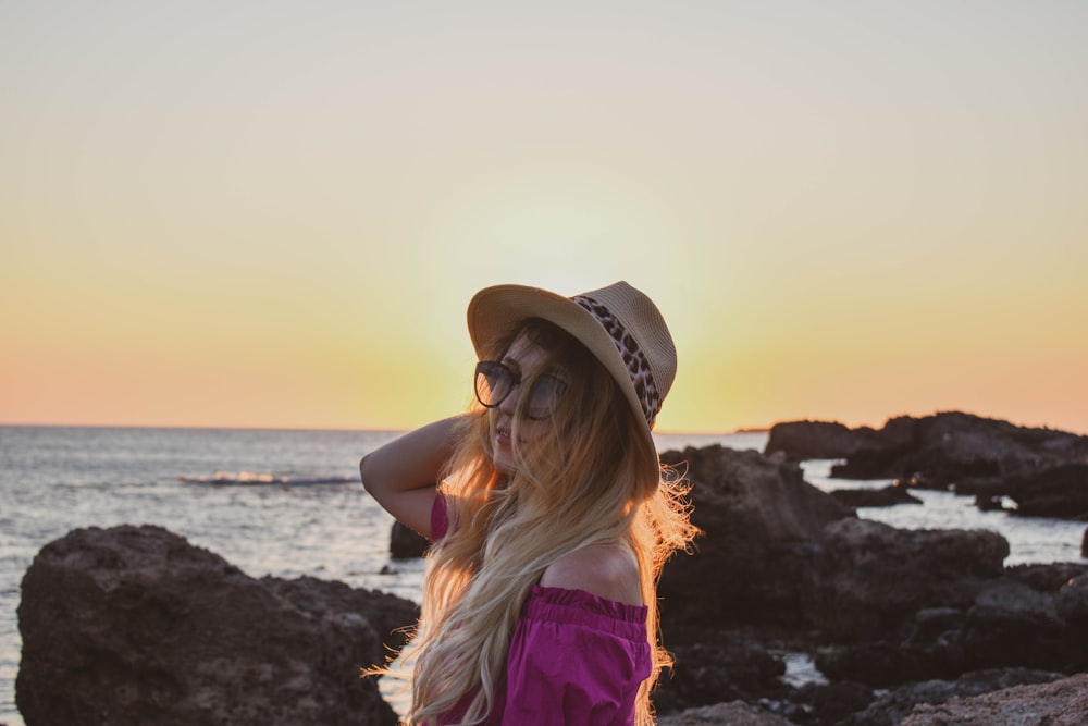 a woman wearing a hat and sunglasses standing on rocks near the ocean