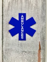 A blue asterisk-shaped sign with the word 'AMBULANCES' written vertically, mounted on a textured, weathered concrete wall with visible cracks and stains.