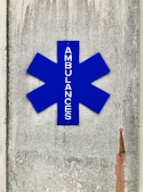 A blue asterisk-shaped sign with the word 'AMBULANCES' written vertically, mounted on a textured, weathered concrete wall with visible cracks and stains.