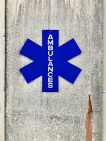 A blue asterisk-shaped sign with the word 'AMBULANCES' written vertically, mounted on a textured, weathered concrete wall with visible cracks and stains.