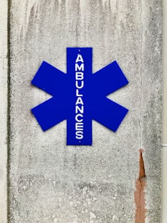 A blue asterisk-shaped sign with the word 'AMBULANCES' written vertically, mounted on a textured, weathered concrete wall with visible cracks and stains.