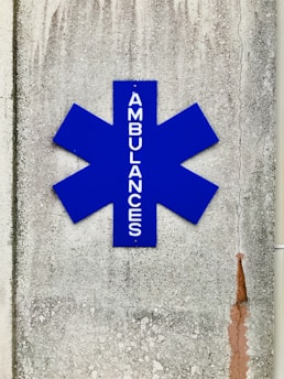 A blue asterisk-shaped sign with the word 'AMBULANCES' written vertically, mounted on a textured, weathered concrete wall with visible cracks and stains.