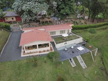 An aerial view of a modern house with a red roof surrounded by lush greenery. The house features a patio with an umbrella, outdoor furniture, and a hot tub. A driveway leads up to the house, and there are a few scattered trees and bushes around the property.