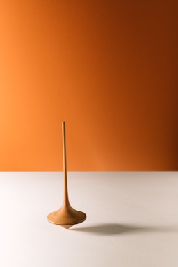 A spinning top with a sleek, elongated design stands upright on a smooth, light-colored surface. The background is a warm, solid orange color that complements the top's earthy tones.