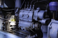 The image depicts a close-up view of industrial machinery, highlighting a section of an engine with various components, including bolts, pipes, and a small glass container with a yellowish liquid. The machinery appears to be metallic with a weathered surface, suggesting regular use.