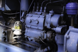 The image depicts a close-up view of industrial machinery, highlighting a section of an engine with various components, including bolts, pipes, and a small glass container with a yellowish liquid. The machinery appears to be metallic with a weathered surface, suggesting regular use.