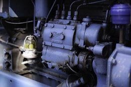The image depicts a close-up view of industrial machinery, highlighting a section of an engine with various components, including bolts, pipes, and a small glass container with a yellowish liquid. The machinery appears to be metallic with a weathered surface, suggesting regular use.