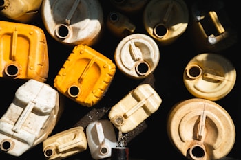 A collection of used, stacked plastic jerrycans is arranged with varying orientations. The containers are mostly yellow and white, showing signs of wear and exposure. The sunlight casts shadows and highlights across their surfaces, creating an intricate pattern of light and dark.