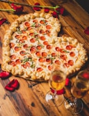 A heart-shaped pizza with a thick, braided crust topped with pepperoni and basil. It sits on a wooden table surrounded by scattered red rose petals. Two flutes of champagne are positioned nearby, reflecting a romantic setting.
