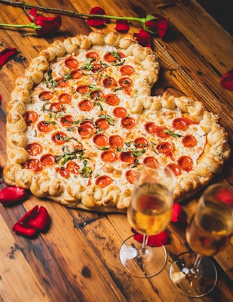 A heart-shaped pizza with a thick, braided crust topped with pepperoni and basil. It sits on a wooden table surrounded by scattered red rose petals. Two flutes of champagne are positioned nearby, reflecting a romantic setting.