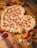 A heart-shaped pizza with a thick, braided crust topped with pepperoni and basil. It sits on a wooden table surrounded by scattered red rose petals. Two flutes of champagne are positioned nearby, reflecting a romantic setting.