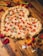 A heart-shaped pizza with a thick, braided crust topped with pepperoni and basil. It sits on a wooden table surrounded by scattered red rose petals. Two flutes of champagne are positioned nearby, reflecting a romantic setting.