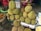 A collection of durians piled up on a stand at a market. Surrounding the durians are other fruits, including rambutans and dark purple mangosteens. Two people are seated in the background, with one wearing a bright, colorful outfit.