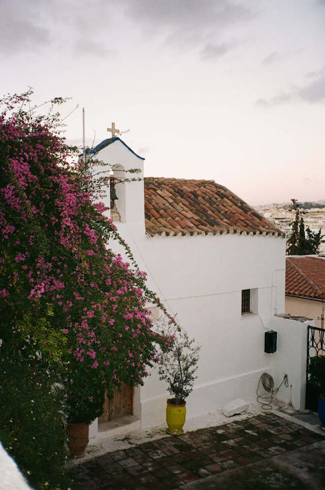 A small whitewashed chapel with a bell tower and blue cross, framed by magenta bougainvillea, overlooking the rooftops of Athens below.