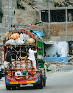 A colorfully decorated truck is heavily loaded with various goods secured by ropes. Two men are hanging onto the back of the truck as it travels down a road. The background consists of unfinished buildings and rubble, suggesting a construction area or an unfinished urban environment.