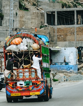 A colorfully decorated truck is heavily loaded with various goods secured by ropes. Two men are hanging onto the back of the truck as it travels down a road. The background consists of unfinished buildings and rubble, suggesting a construction area or an unfinished urban environment.