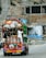 A colorfully decorated truck is heavily loaded with various goods secured by ropes. Two men are hanging onto the back of the truck as it travels down a road. The background consists of unfinished buildings and rubble, suggesting a construction area or an unfinished urban environment.
