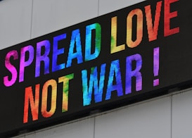 A digital sign displaying the phrase 'Spread Love, Not War!' in vibrant, colorful letters. The text is arranged in multiple colors against a white background, creating a bright and positive visual effect.