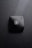 A black compact makeup case rests on a surface covered with water droplets. The compact case is sleek and has a logo with intertwined letters in the center, creating a sophisticated and luxurious impression.