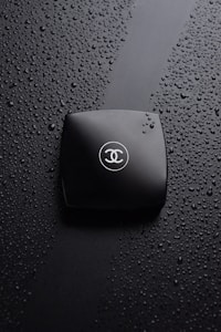 A black compact makeup case rests on a surface covered with water droplets. The compact case is sleek and has a logo with intertwined letters in the center, creating a sophisticated and luxurious impression.