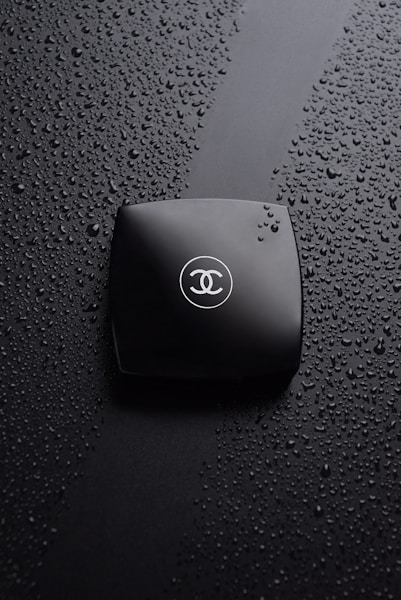A black compact makeup case rests on a surface covered with water droplets. The compact case is sleek and has a logo with intertwined letters in the center, creating a sophisticated and luxurious impression.