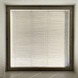 A large window with closed horizontal blinds, allowing minimal light through. The blinds have a neutral beige tone, and the window is set in a recessed wall, casting subtle shadows on the sill.