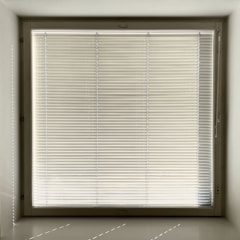 A large window with closed horizontal blinds, allowing minimal light through. The blinds have a neutral beige tone, and the window is set in a recessed wall, casting subtle shadows on the sill.