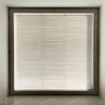 A large window with closed horizontal blinds, allowing minimal light through. The blinds have a neutral beige tone, and the window is set in a recessed wall, casting subtle shadows on the sill.