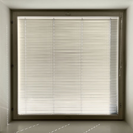 A large window with closed horizontal blinds, allowing minimal light through. The blinds have a neutral beige tone, and the window is set in a recessed wall, casting subtle shadows on the sill.