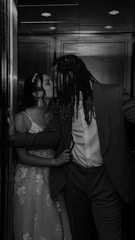 A black and white photograph of a couple sharing a kiss in an elevator. The woman is wearing a lace dress and has her hair in an updo with a headpiece. The man is dressed in a suit and has long dreadlocks. The lighting creates a romantic and intimate ambiance.