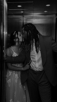 A black and white photograph of a couple sharing a kiss in an elevator. The woman is wearing a lace dress and has her hair in an updo with a headpiece. The man is dressed in a suit and has long dreadlocks. The lighting creates a romantic and intimate ambiance.