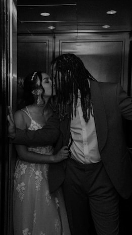 A black and white photograph of a couple sharing a kiss in an elevator. The woman is wearing a lace dress and has her hair in an updo with a headpiece. The man is dressed in a suit and has long dreadlocks. The lighting creates a romantic and intimate ambiance.
