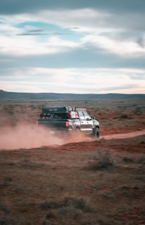A black pickup truck drives off-road through a dusty landscape, leaving a trail of dust behind. The vehicle is equipped with gear on its roof rack, suggesting an adventurous journey. The vast, open terrain is barren with patches of dry grass, and the sky above is filled with clouds.