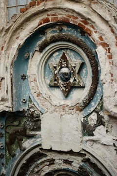 A detailed and deteriorated architectural relief featuring a Star of David at the center, surrounded by ornate designs and brickwork. The background shows peeling paint, suggesting age and weathering. Below the star, there is a sculpted lion and an open book motif, both blending with the eroded surface.