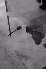A reflective puddle on the pavement shows the silhouette of a streetlamp and part of a building. Nearby, two feet wearing shoes, one in motion, are visible, suggesting a person walking.