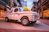 A vintage white pickup truck is parked in an urban setting, adorned with vibrant purple and yellow flowers in its bed. The truck displays the words 'Pearl in full bloom' on its side. It is situated on a paved pathway flanked by modern buildings, one of which has an illuminated sign that reads 'ARIAT'. The lighting suggests either early evening or morning, with a warm ambiance created by street lights.