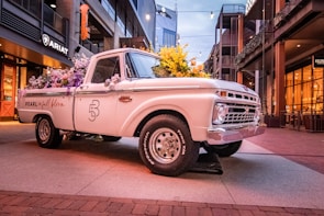 A vintage white pickup truck is parked in an urban setting, adorned with vibrant purple and yellow flowers in its bed. The truck displays the words 'Pearl in full bloom' on its side. It is situated on a paved pathway flanked by modern buildings, one of which has an illuminated sign that reads 'ARIAT'. The lighting suggests either early evening or morning, with a warm ambiance created by street lights.
