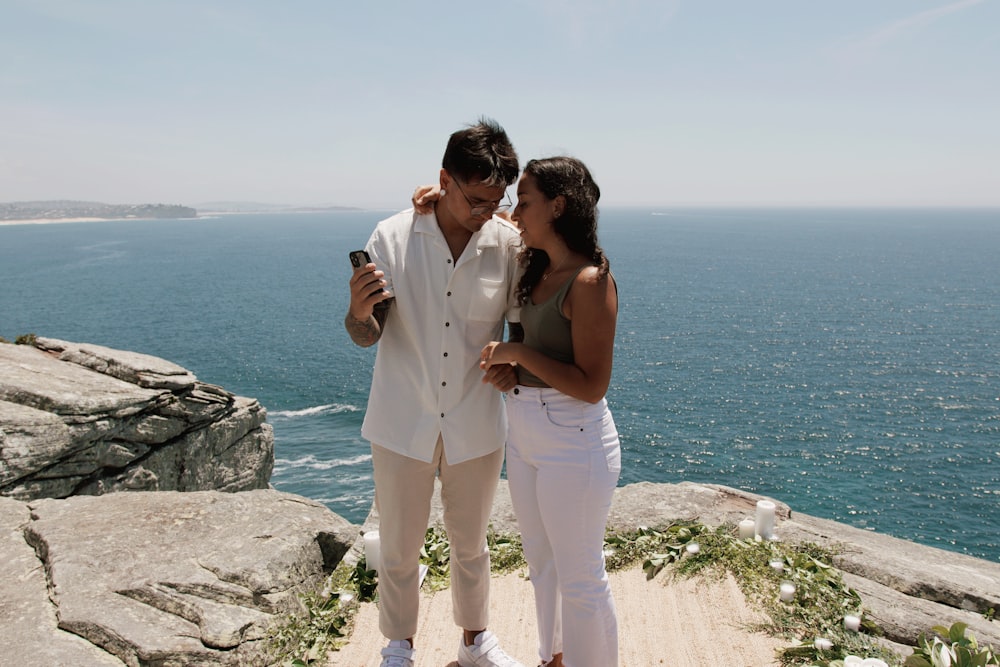 a man and a woman standing on top of a cliff next to the ocean