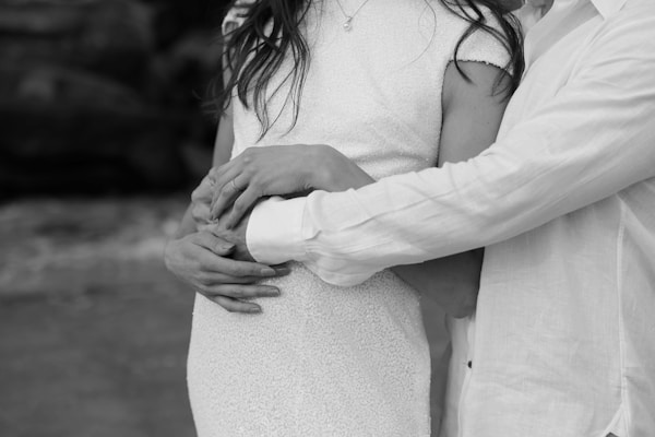 A couple is embracing tenderly, with their hands gently placed on each other's arms and midsection. The focus is on their intertwined hands and the intimacy of their pose. They are wearing light-colored clothing, set against a blurred background.