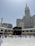 A large gathering of people surrounding the Kaaba in Mecca, with the Abraj Al-Bait Clock Tower and other skyscrapers in the background. The courtyard is spacious and bright, filled with individuals dressed in traditional white garments, denoting a religious pilgrimage.